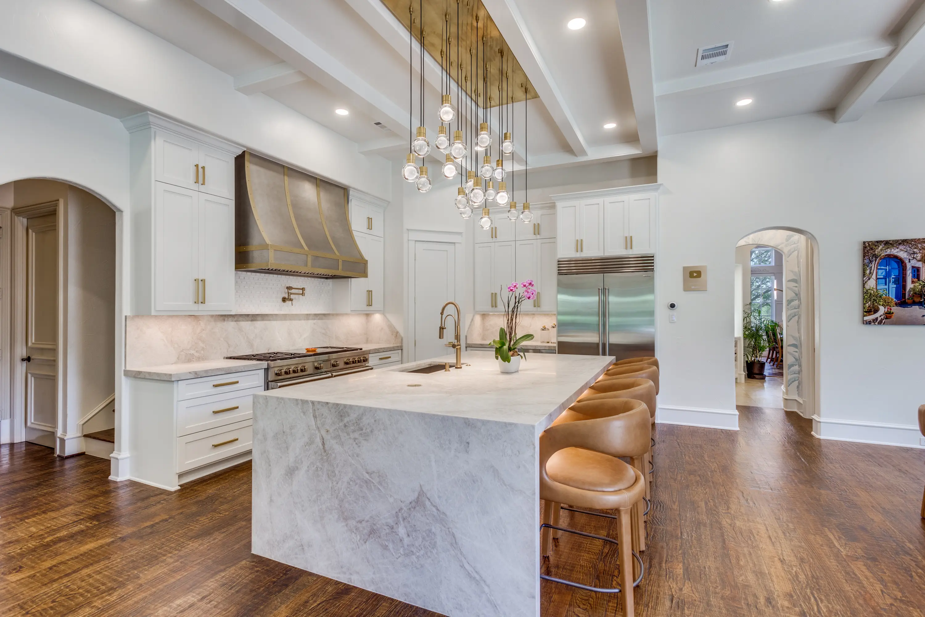 An open kitchen with a waterfall Taj Mahal quartzite island, a custom brass-banded range hood, and a cascading pendant light.