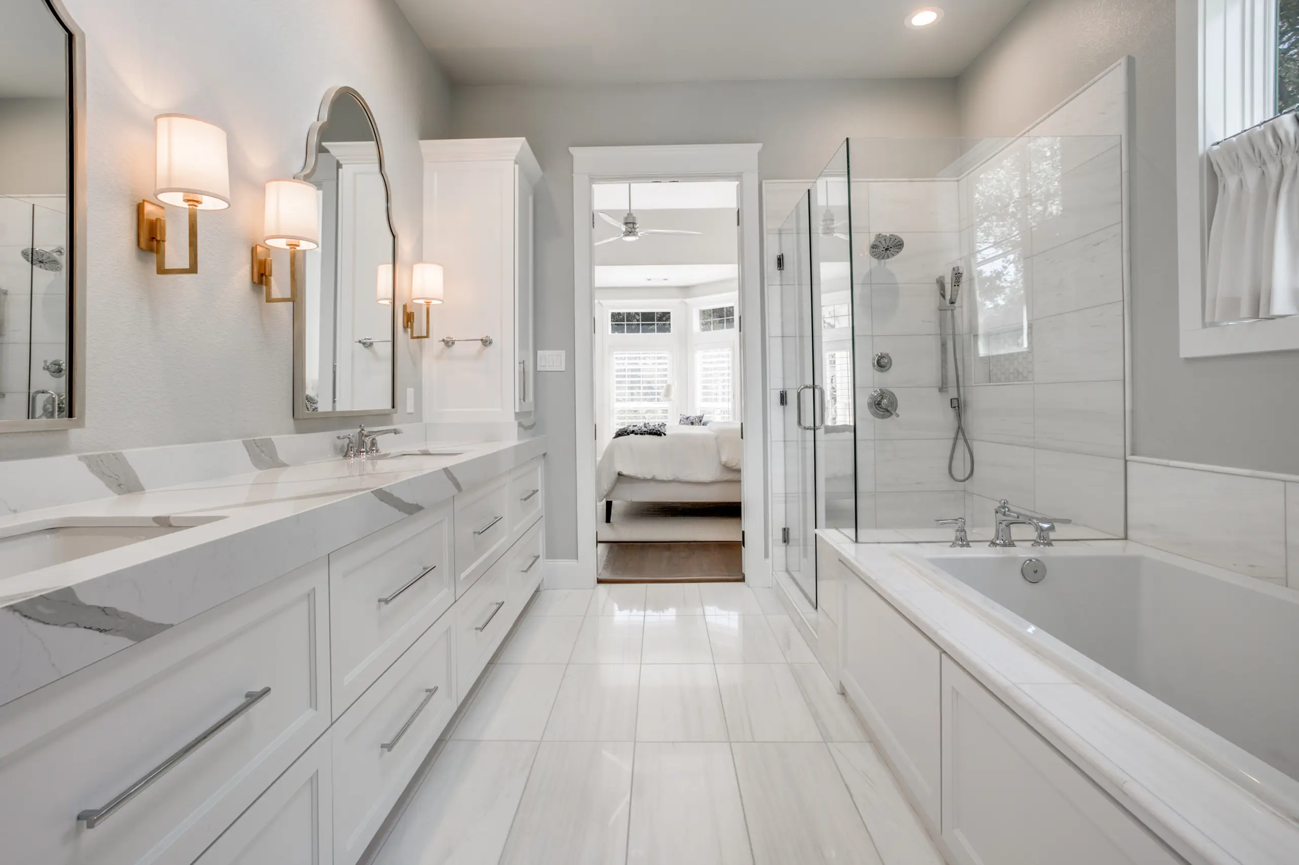 A bright white primary bath with a marble double vanity, brass sconces, glass shower, and a built-in soaking tub.